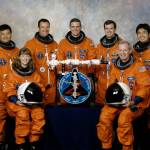 Seven astronauts in orange spacesuits pose in front of US and Japanese flag