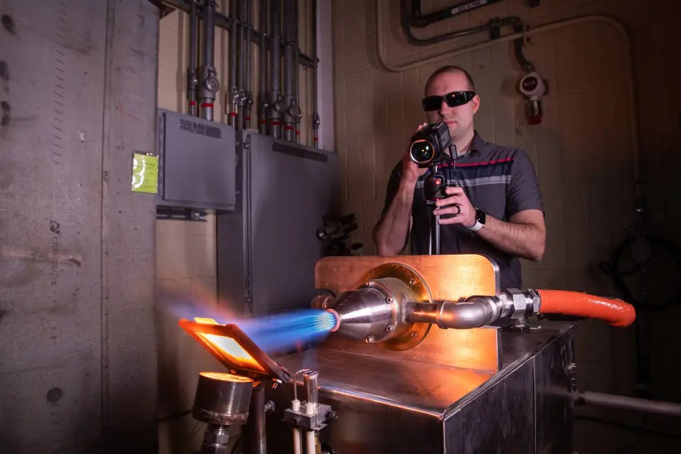 A man working in an industrial laboratory wearing sunglasses points a camera at a silver exhaust port emitting a superhot blue flame that is striki...