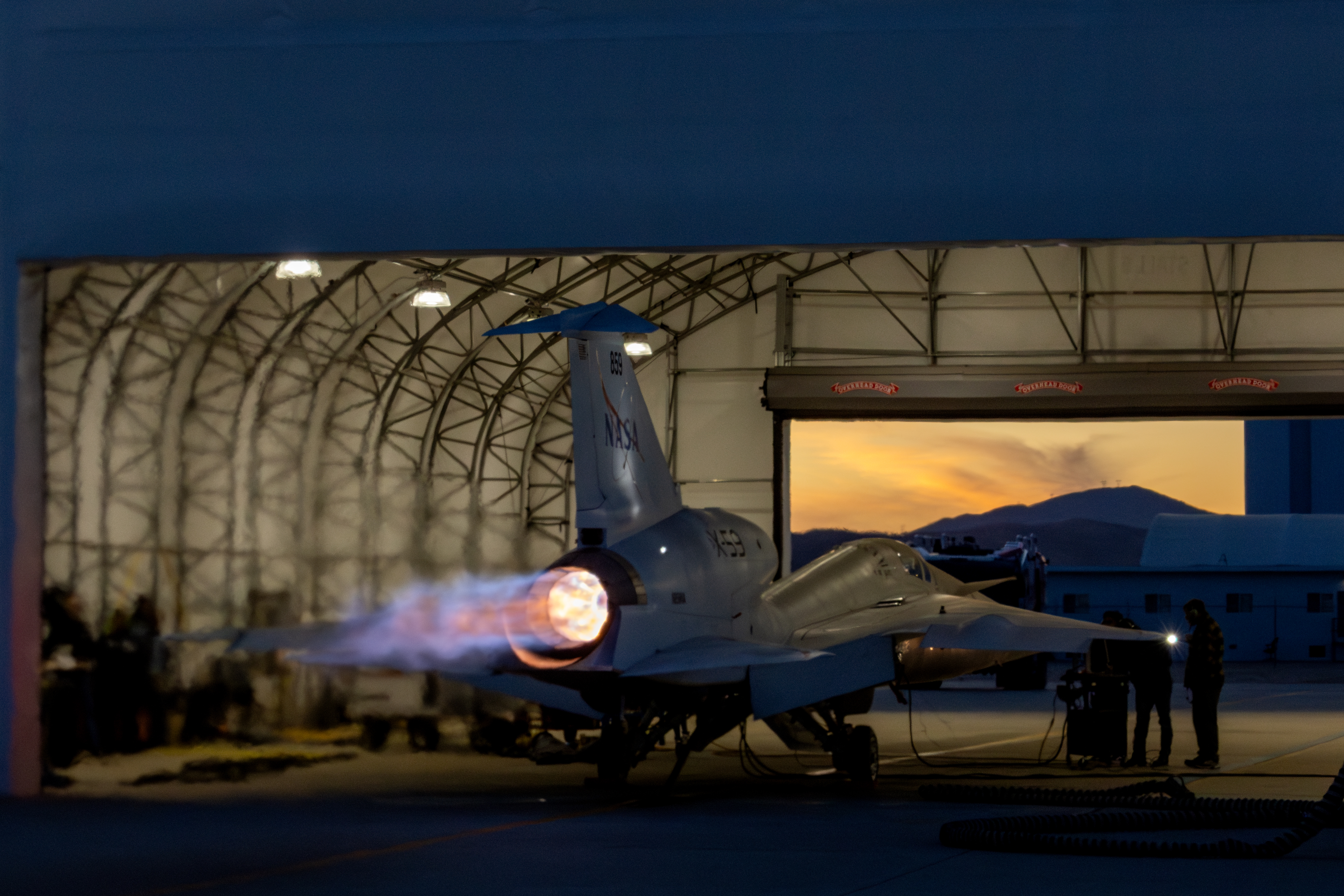 NASA’s X-59 sits partially in a white hangar and emits bright flames with visible shock diamonds from its engine at sunset during maximum afterbu...